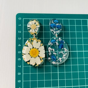 K&ouml;nnte beinhalten: Ein Paar H&auml;ngeohrringe mit klarem Harz und gepressten Blumen. Ein Ohrring zeigt eine wei&szlig;e G&auml;nsebl&uuml;mchen mit goldenen Akzenten und der andere zeigt blaue Blumen.
