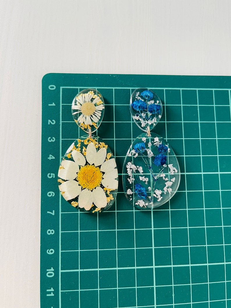 K&ouml;nnte beinhalten: Ein Paar H&auml;ngeohrringe mit klarem Harz und gepressten Blumen. Ein Ohrring zeigt eine wei&szlig;e G&auml;nsebl&uuml;mchen mit goldenen Akzenten und der andere zeigt blaue Blumen.
