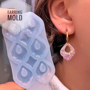 Könnte beinhalten: Eine transparente, hellblaue Ohrringform mit einem sich wiederholenden Tropfendesign. Die Form wird neben einem goldenen Creolen-Ohrring mit einem rosa, glitzernden, tropfenförmigen Anhänger gehalten. Der Text "EARRING MOLD" ist sichtbar.