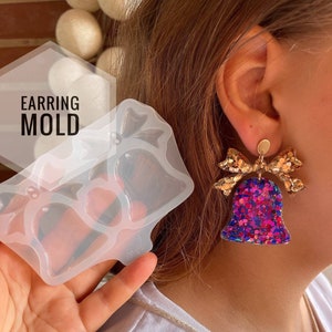 May include: A clear silicone mold for making earrings in the shape of a bell with a bow. The mold is shown next to a pair of finished earrings with a gold bow and a bell covered in iridescent glitter.
