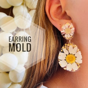 K&ouml;nnte beinhalten: Ein Paar Resin-Ohrringe mit einem gepressten Blumendesign. Die Ohrringe sind rund und zeigen eine wei&szlig;e Margerite mit gelber Mitte. Die Ohrringe h&auml;ngen an einem Ohr einer Person.