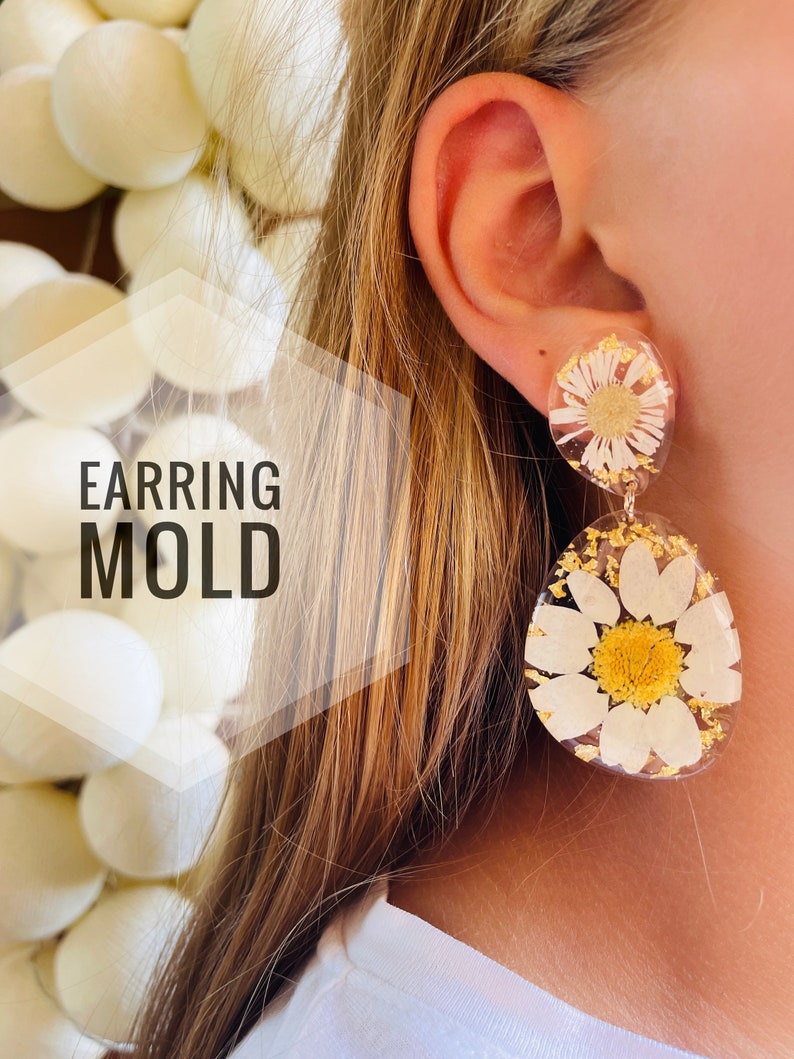 K&ouml;nnte beinhalten: Ein Paar Resin-Ohrringe mit einem gepressten Blumendesign. Die Ohrringe sind rund und zeigen eine wei&szlig;e Margerite mit gelber Mitte. Die Ohrringe h&auml;ngen an einem Ohr einer Person.