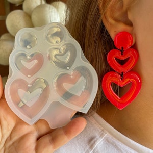 Pode incluir: Um molde de silicone transparente com quatro cavidades em forma de coração. Um par de brincos vermelhos em forma de coração é mostrado no fundo.