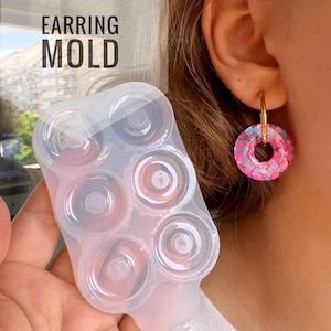 以下が含まれることがあります： イヤリングを作るための、6つの丸い空洞がある透明なシリコンモールド。モールドは手で持たれており、背景にはピンクのグリッターイヤリングのペアが映っています。