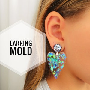 May include: A pair of iridescent heart-shaped earrings with a silver glitter accent. The earrings are made of clear resin with blue and green glitter embedded in the resin. The earrings are hanging from a person's ear.