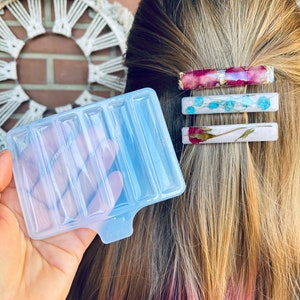 Puede incluir: Un molde de silicona transparente con seis cavidades rectangulares para hacer pinzas para el pelo de resina. Se muestran tres pinzas para el pelo de resina acabadas con flores secas y detalles dorados en el fondo.