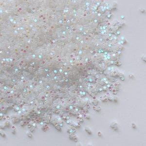 Puede incluir: Primer plano de un montón de purpurina blanca e iridiscente. Las partículas de purpurina son pequeñas, rectangulares y reflejan un arcoíris de colores, incluyendo rosa, azul y amarillo. La purpurina está esparcida sobre una superficie blanca.
