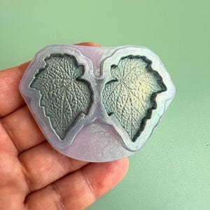 May include: A translucent silicone mold featuring two recessed leaf-shaped cavities with intricate vein detailing. The mold has a pearlescent, light purple tint, while the interior leaf impressions display a shimmering, metallic green finish.