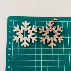 Op de afbeelding: Twee oorbelletjes in de vorm van een sneeuwvlok, een is wit en de andere is roze met glitter. De oorbelletjes liggen op een groene snijmat.