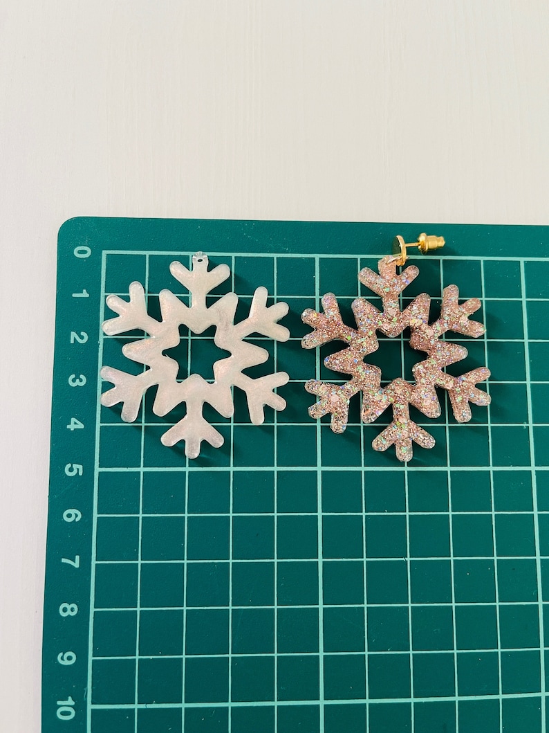 Op de afbeelding: Twee oorbelletjes in de vorm van een sneeuwvlok, een is wit en de andere is roze met glitter. De oorbelletjes liggen op een groene snijmat.