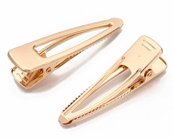 Op de afbeelding: Twee gouden metalen haarclips. De clips zijn lang en smal met een getextureerde grip.