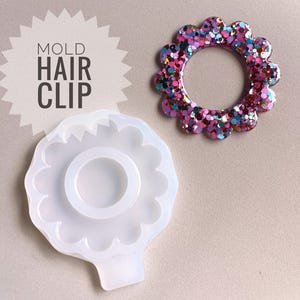 Könnte beinhalten: Eine weiße Silikonform zum Herstellen einer Haarklammer. Die Form ist blumenförmig mit einer runden Mitte. Die Form wird neben einer fertigen Haarklammer gezeigt, die mit rosa, lila und goldenen Glitzer hergestellt wurde.