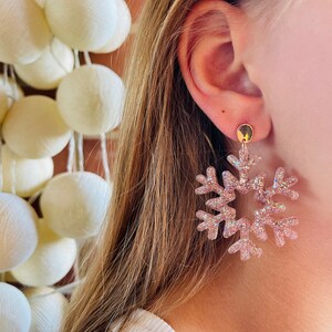Op de afbeelding: Een paar roze glitter sneeuwvlok oorbellen. De oorbellen hangen en hebben een gouden oorbel.