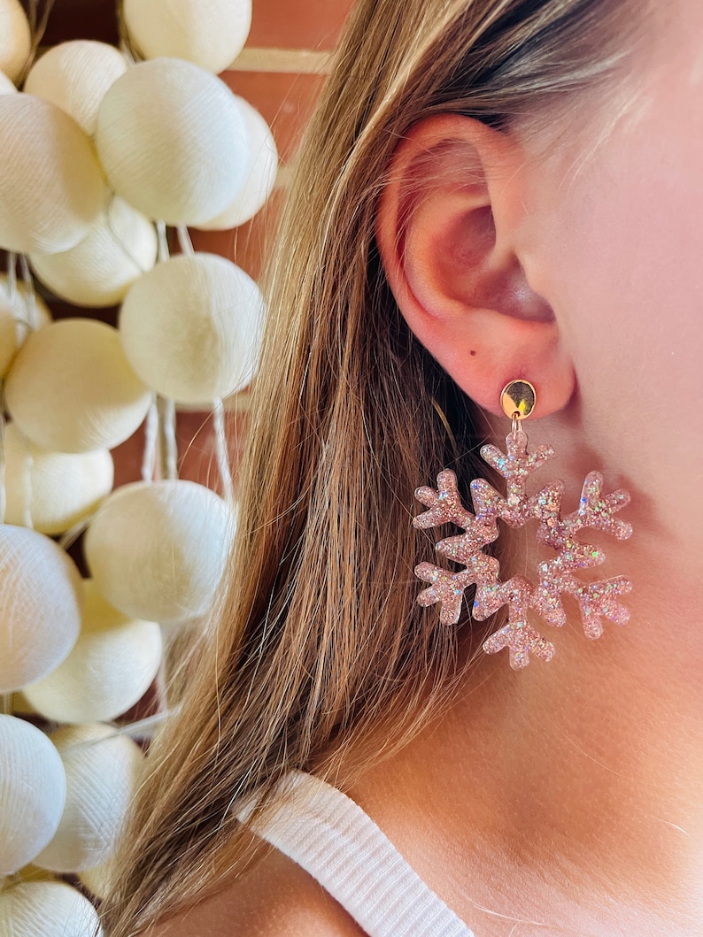Op de afbeelding: Een paar roze glitter sneeuwvlok oorbellen. De oorbellen hangen en hebben een gouden oorbel.