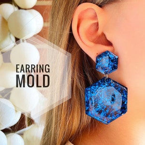 May include: A pair of blue hexagonal resin earrings with a glittery finish. The earrings are shown on a model's ear.
