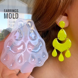 Puede incluir: Un molde de aretes de silicona transparente con el texto "EARRINGS MOLD" y un par de aretes de araña de color amarillo neón. Los aretes tienen un diseño de tres niveles con una parte superior curva y colgantes en forma de lágrima. El molde está diseñado.