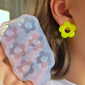 Può includere: Uno stampo in silicone trasparente con sei cavità a forma di fiore è tenuto vicino all'orecchio di una persona, adornato da un orecchino giallo brillante a forma di fiore. Lo stampo serve per realizzare gioielli in resina o oggetti decorativi.