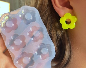 Può includere: Uno stampo in silicone trasparente con sei cavità a forma di fiore è tenuto vicino all'orecchio di una persona, adornato da un orecchino giallo brillante a forma di fiore. Lo stampo serve per realizzare gioielli in resina o oggetti decorativi.