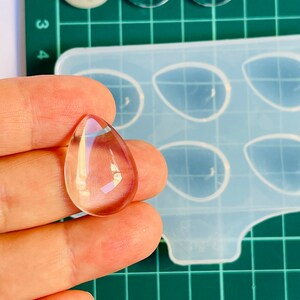 Puede incluir: Un colgante de resina transparente en forma de lágrima con un ligero tono rosado. El colgante está sobre una mano, y en el fondo hay un molde de silicona con seis cavidades en forma de lágrima.