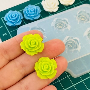 May include: Two bright green resin rose flower cabochons, ideal for jewellery making or crafting. The roses are shown on a hand, with a silicone mould in the background.