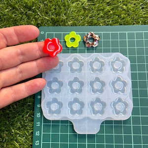 以下が含まれることがあります： 透明なシリコンモールドで、12個の花の形をした空洞があります。モールドは緑色のカッティングマットの上に置かれています。モールドの隣には、花の形をしたレジン製のものが3つあります。1つは赤、1つは黄色、もう1つは茶色と白のグリッターです。