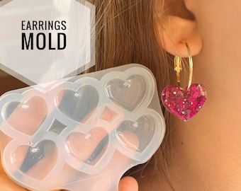 Può includere: Uno stampo per orecchini in silicone trasparente con cavità a forma di cuore è tenuto vicino a un orecchino a cerchio dorato con un ciondolo a forma di cuore rosa glitterato. Il testo "EARRINGS MOLD" è in un esagono bianco.