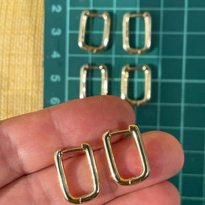 Pode incluir: Brincos de argola retangulares dourados. Os brincos têm um design simples e moderno com um acabamento polido. Vários pares de brincos são visíveis, alguns dispostos sobre uma base de corte verde.