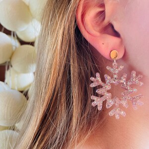 Op de afbeelding: Een paar sneeuwvlok oorbellen met een roze glitterafwerking. De oorbellen zijn gemaakt van helder plastic en hebben een gouden post.