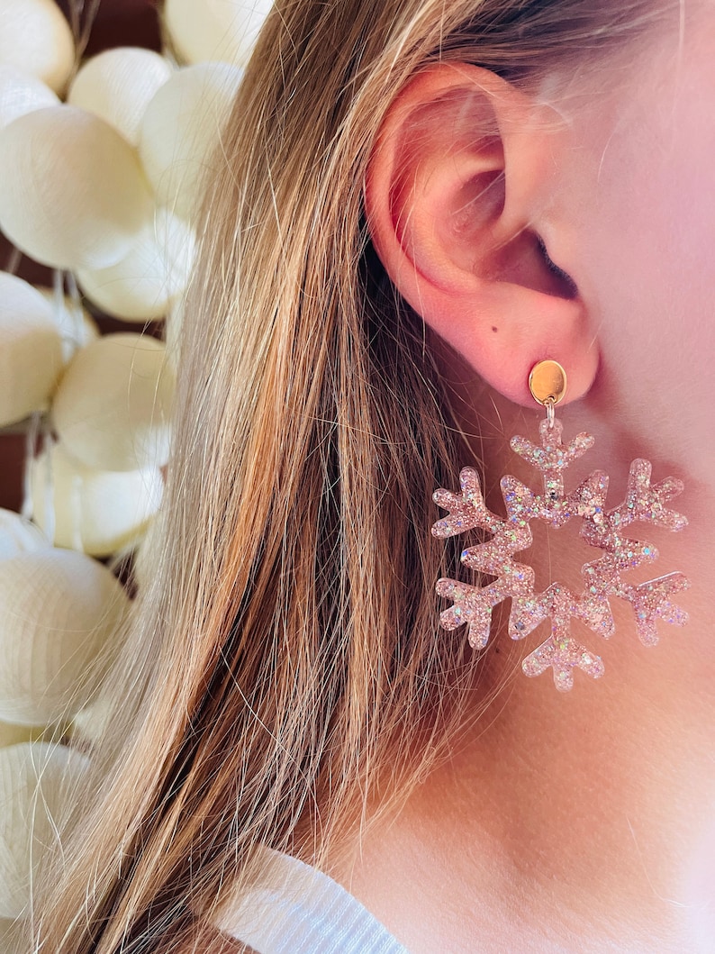 Op de afbeelding: Een paar sneeuwvlok oorbellen met een roze glitterafwerking. De oorbellen zijn gemaakt van helder plastic en hebben een gouden post.