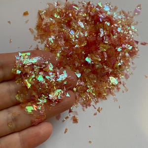 May include: Iridescent orange and gold flakes scattered on a white surface. Some flakes are held in a hand. The flakes have a shimmering, reflective quality, with hints of green and blue.