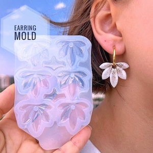 Puede incluir: Un molde de silicona translúcido para pendientes con seis cavidades en forma de hoja y el texto EARRING MOLD impreso en la esquina superior izquierda. Junto al molde hay un pendiente de aro dorado con un colgante blanco y dorado con purpurina.