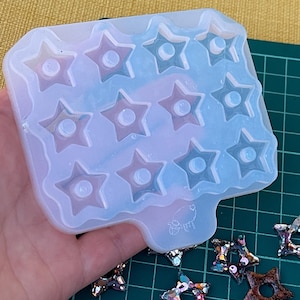 May include: A clear silicone mold with star-shaped cavities, each with a central hole. The mold is held in a hand, with several finished star-shaped resin pieces visible nearby. The mold is on a green cutting mat.
