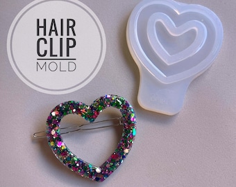 Può includere: Uno stampo in silicone trasparente a forma di cuore con un cuore più piccolo all'interno. Lo stampo viene utilizzato per creare fermagli per capelli in resina. Il testo "HAIR CLIP MOLD" è visibile nell'immagine.