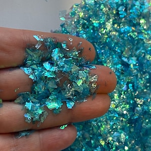 May include: Close-up of iridescent blue and green glitter flakes. The glitter is in the shape of small, irregular squares and is scattered across a surface. The glitter has a reflective quality, catching the light and creating a shimmering effect.