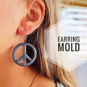 Può includere: Un paio di orecchini a forma di segno della pace con una finitura blu e viola scintillante. Gli orecchini sono appesi all'orecchio di una persona. Il testo "EARRING MOLD" è visibile nell'immagine.