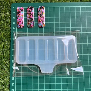 Könnte beinhalten: Eine transparente Silikonform mit sechs rechteckigen Vertiefungen zum Herstellen von Kunsthandwerk aus Harz. Die Form hat einen Griff und ist in einer transparenten Plastiktüte verpackt. Drei kleine rechteckige Harzstücke mit buntem Glitzer sind neben der Form abgebildet.