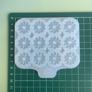 Peut inclure: Un moule en silicone translucide comportant douze cavités en forme de fleur disposées en grille, avec de petits accents en forme de losange entre elles. Le moule est posé sur un tapis de découpe vert avec des lignes blanches, mesurant 10 cm.