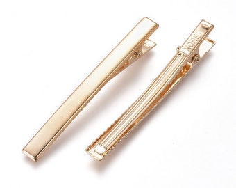 Op de afbeelding: Twee goudkleurige metalen haarclips. Eén clip is effen, terwijl de andere een getextureerd interieur heeft en het woord "KOREA" erop gestempeld. Beide clips hebben een eenvoudig, rechthoekig ontwerp met een scharnierende sluiting.