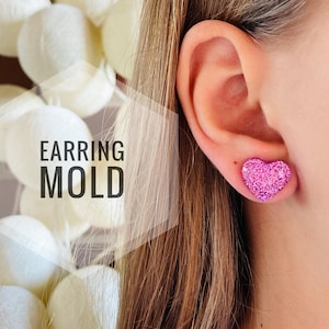 Puede incluir: Un par de pendientes de corazón con purpurina rosa. Los pendientes se muestran en el lóbulo de la oreja de una persona. El texto "EARRING MOLD" es visible en la imagen.