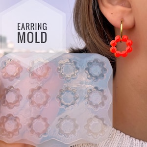 Puede incluir: Un molde de pendientes de silicona transparente con múltiples cavidades en forma de flor se sostiene frente a una persona. Un pendiente rojo en forma de flor cuelga de un pendiente de aro dorado en la oreja de la persona. El texto "EARRING MOLD" está en una caja blanca.