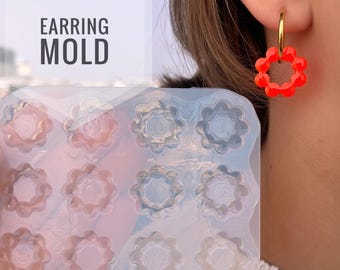 Può includere: Uno stampo per orecchini in silicone trasparente con più cavità a forma di fiore è tenuto davanti a una persona. Un orecchino rosso a forma di fiore pende da un orecchino a cerchio dorato sull'orecchio della persona. Il testo "EARRING MOLD" è in una scatola bianca.