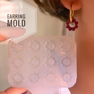 Könnte beinhalten: Eine transparente Silikonform mit 12 blütenförmigen Ohrring-Vertiefungen. Die Form wird mit einem fertigen Ohrring in Blütenform gezeigt, der aus rotem Harz hergestellt wurde.