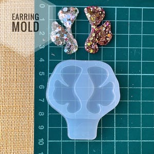 May include: A clear, flexible earring mold with a butterfly design, alongside two finished glitter earrings. One earring is silver, the other is a mix of gold and brown. The text "EARRING MOLD" is visible.