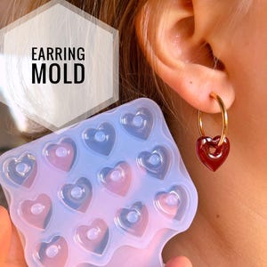 Puede incluir: Un molde de silicona transparente con 12 cavidades en forma de corazón para hacer pendientes. El molde se está sosteniendo frente a la oreja de una persona que lleva un par de pendientes en forma de corazón rojo.