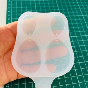 K&ouml;nnte beinhalten: Eine transparente Silikonform mit zwei ovalen Vertiefungen zum Herstellen von Ohrringen. Die Form hat einen kleinen Griff mit einem Schweinemuster.