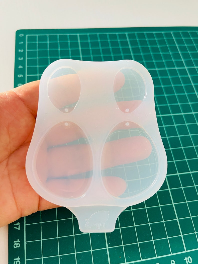 K&ouml;nnte beinhalten: Eine transparente Silikonform mit zwei ovalen Vertiefungen zum Herstellen von Ohrringen. Die Form hat einen kleinen Griff mit einem Schweinemuster.