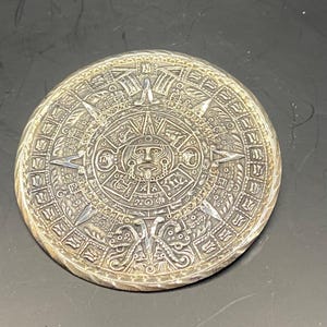 May include: A silver pendant featuring a detailed depiction of the Aztec calendar, also known as the Sun Stone. The intricate design includes a central face surrounded by concentric circles of glyphs and symbols.