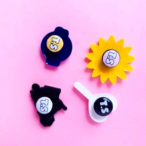 May include: Four different pins with a yellow, blue, black, and white background. Each pin has a white circle with the text "T.S." in black. The pins are on a pink background.