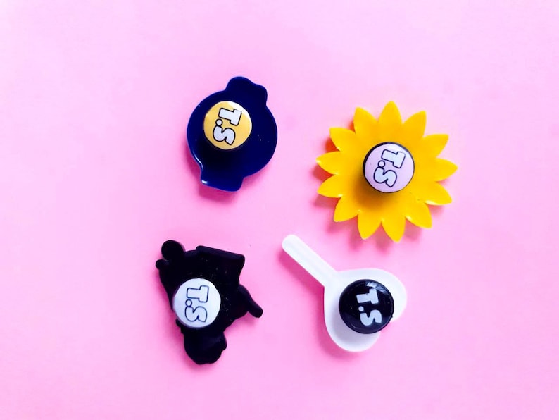 May include: Four different pins with a yellow, blue, black, and white background. Each pin has a white circle with the text "T.S." in black. The pins are on a pink background.