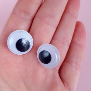May include: Two white plastic googly eyes with black pupils. The eyes are on a person's hand.